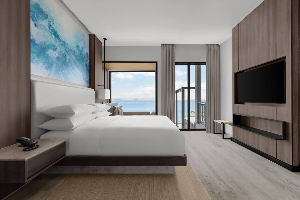 A modern hotel room with a large bed, a flat-screen TV, a nightstand, and a view of the ocean.