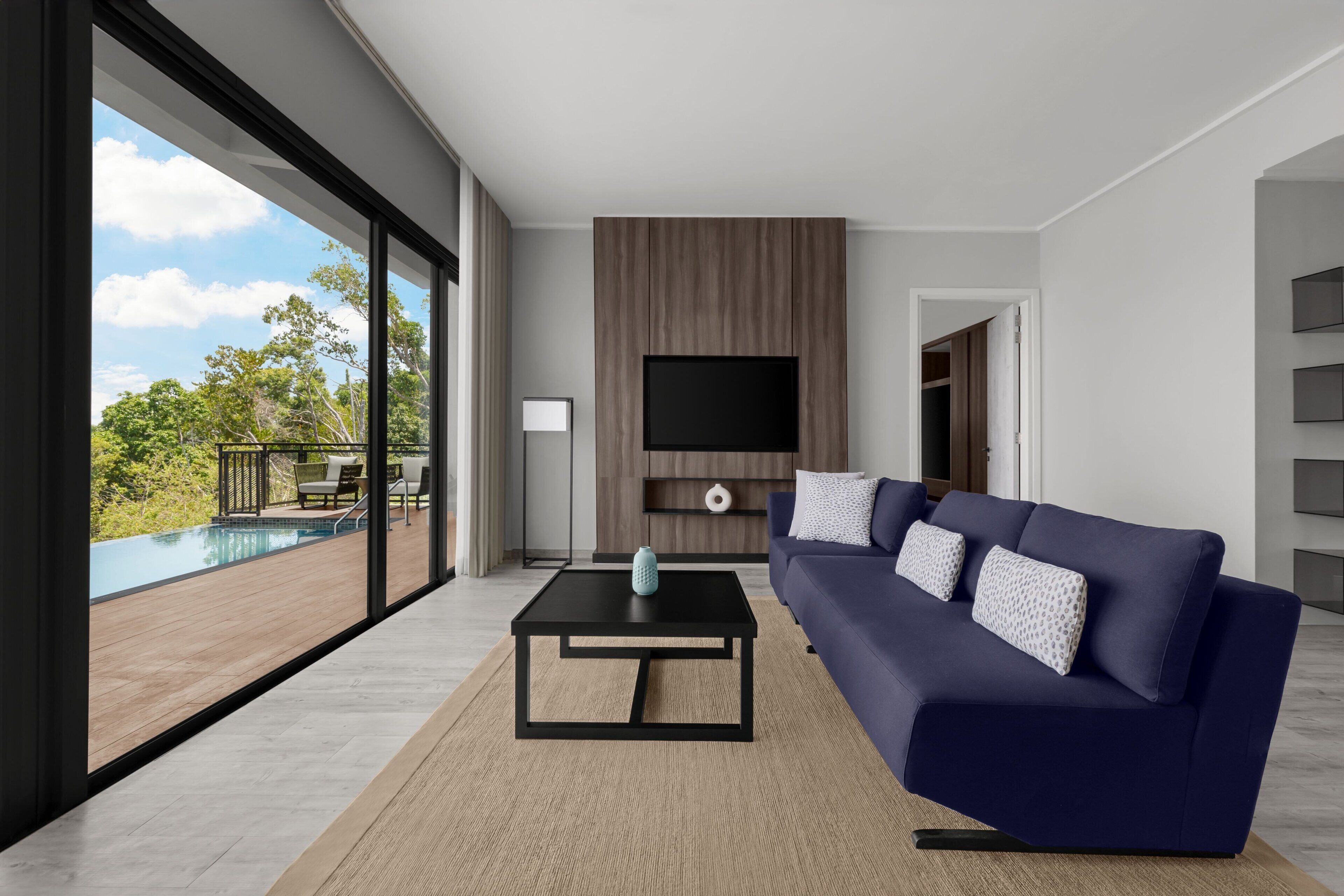 A modern living room with a large TV, a dark blue sofa, and a view of a pool and greenery outside.