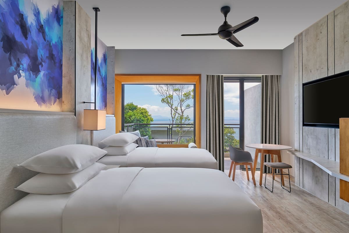 A hotel room with two beds, a TV, a ceiling fan, a balcony with a table and chairs, and a large window with a view of the outdoors.
