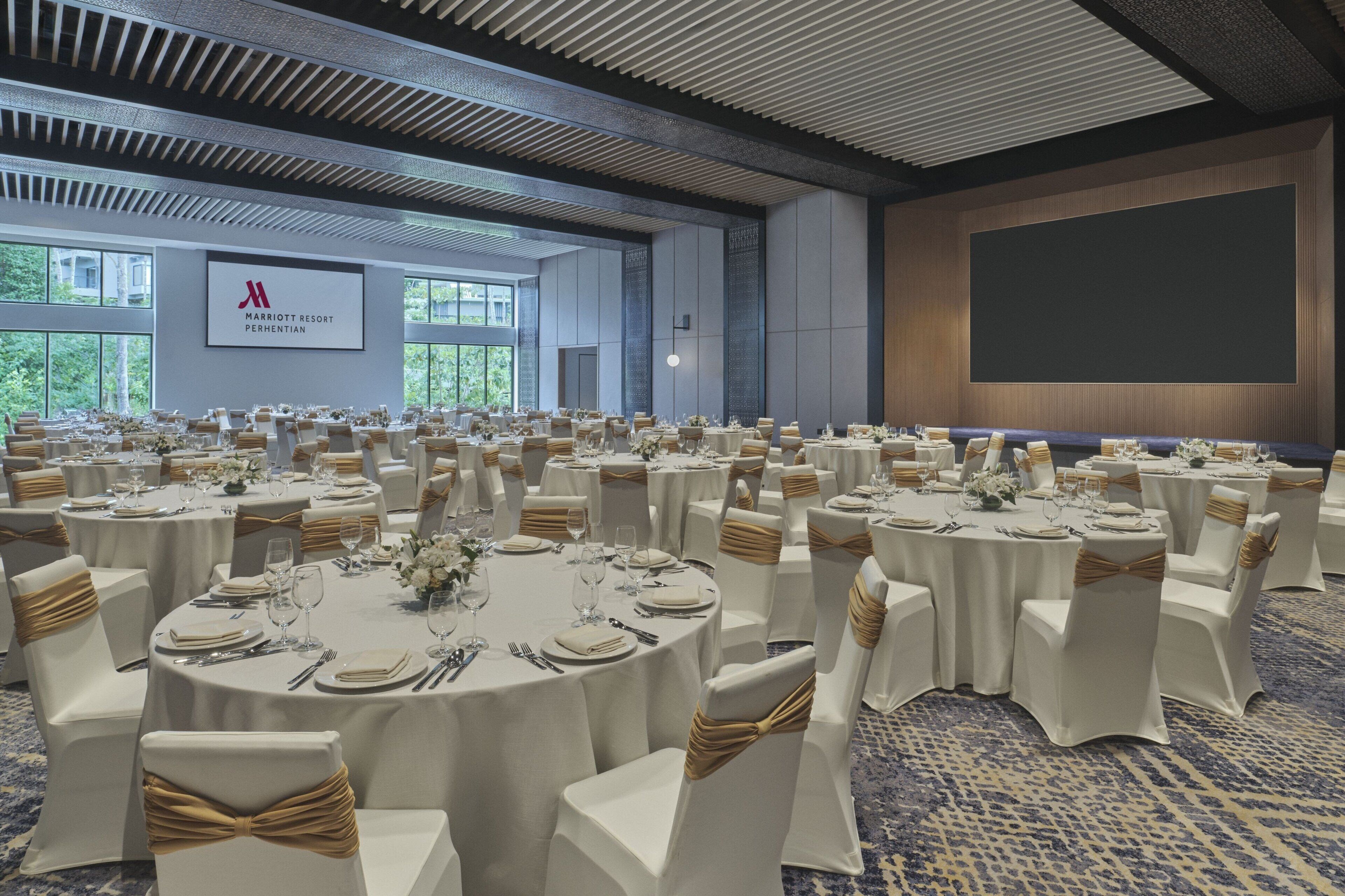 A conference room with round tables, white chairs, and gold sashes. A large screen is mounted on the wall, and the room has a high ceiling with a patterned design.
