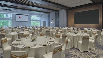A conference room with round tables, white chairs, and gold sashes. A large screen is mounted on the wall, and the room has a high ceiling with a patterned design.