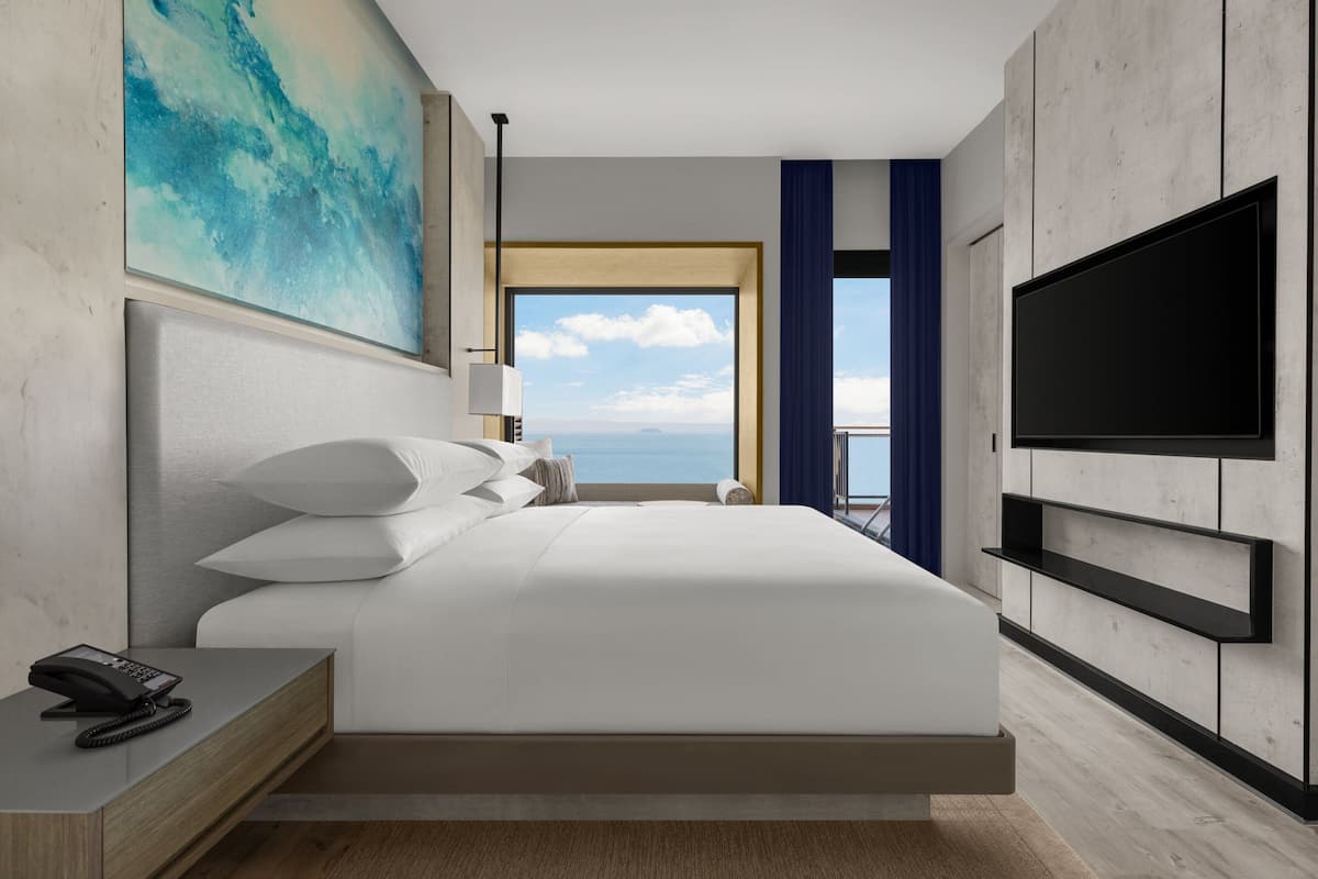 A modern hotel room with a large bed, a flat-screen TV, a bedside phone, and a view of the sea through a large window.