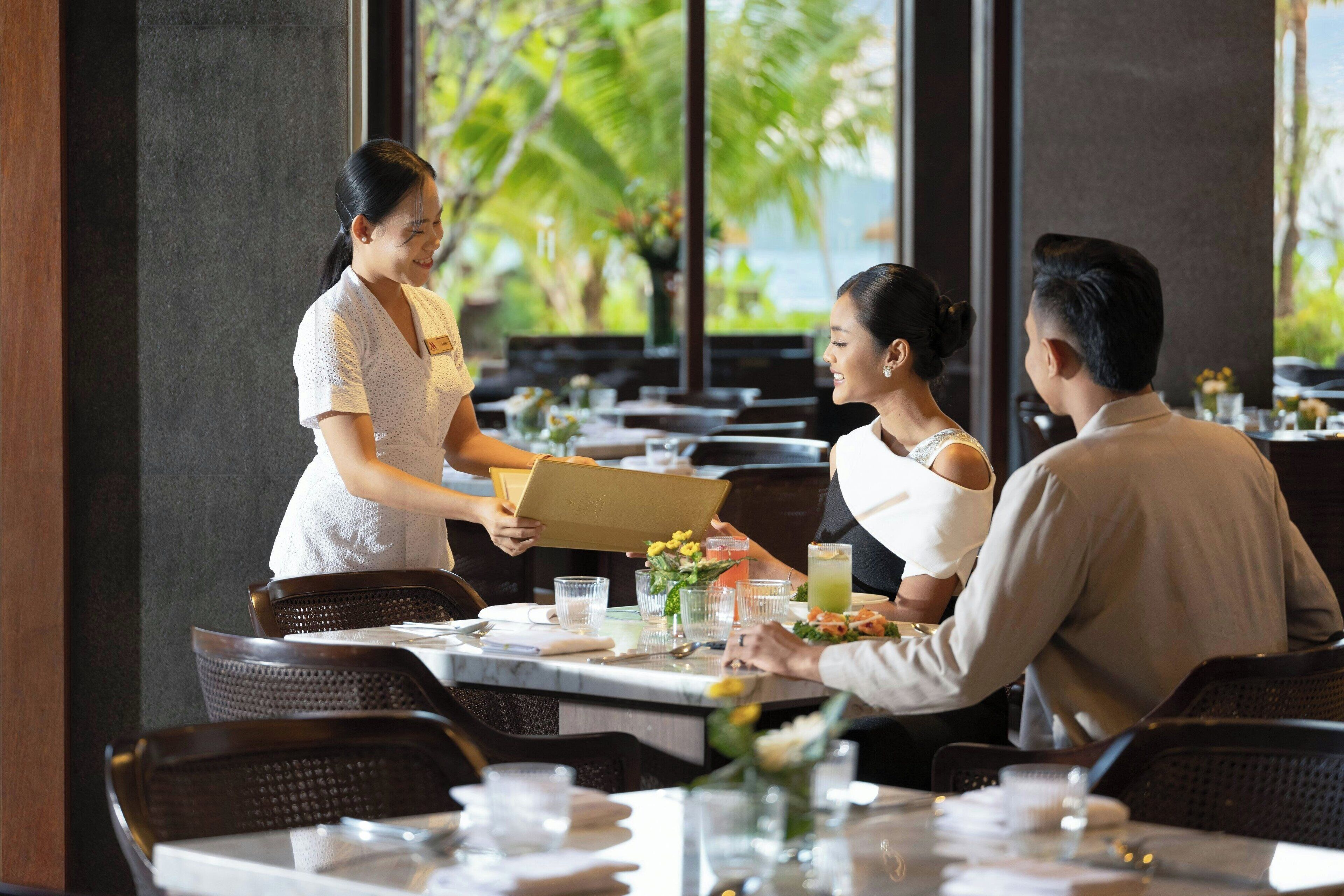 Een restaurant met een ober die een maaltijd serveert aan een stel. Tafels zijn gedekt met glazen, bestek en een vaas met bloemen.