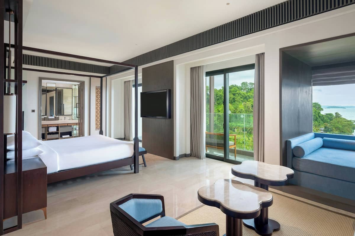 A modern hotel room with a large bed, a flat-screen TV, a balcony with a view of the sea, and a seating area with a table.