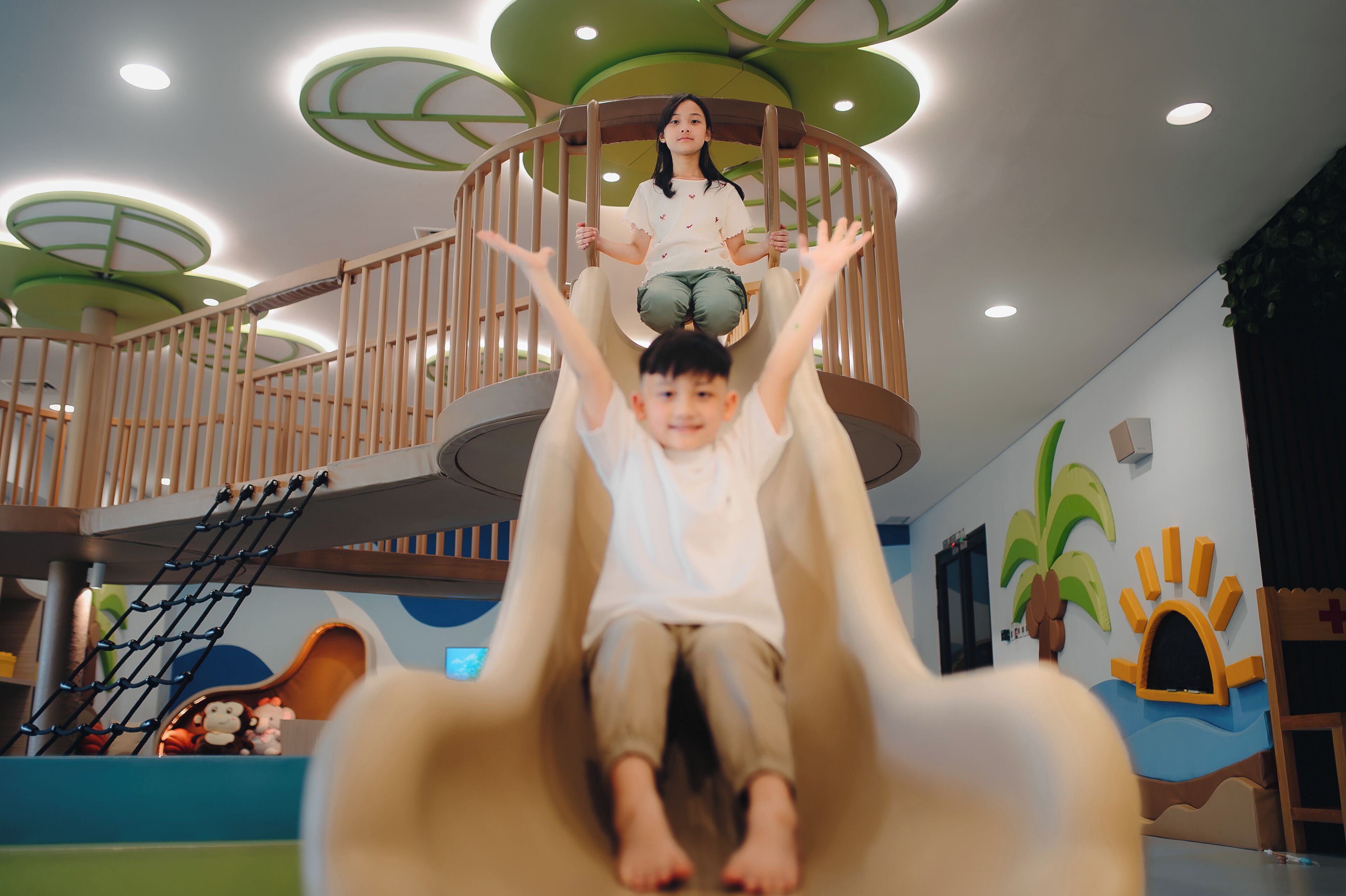 A child sliding down a slide with a wooden railing and a person standing on top.