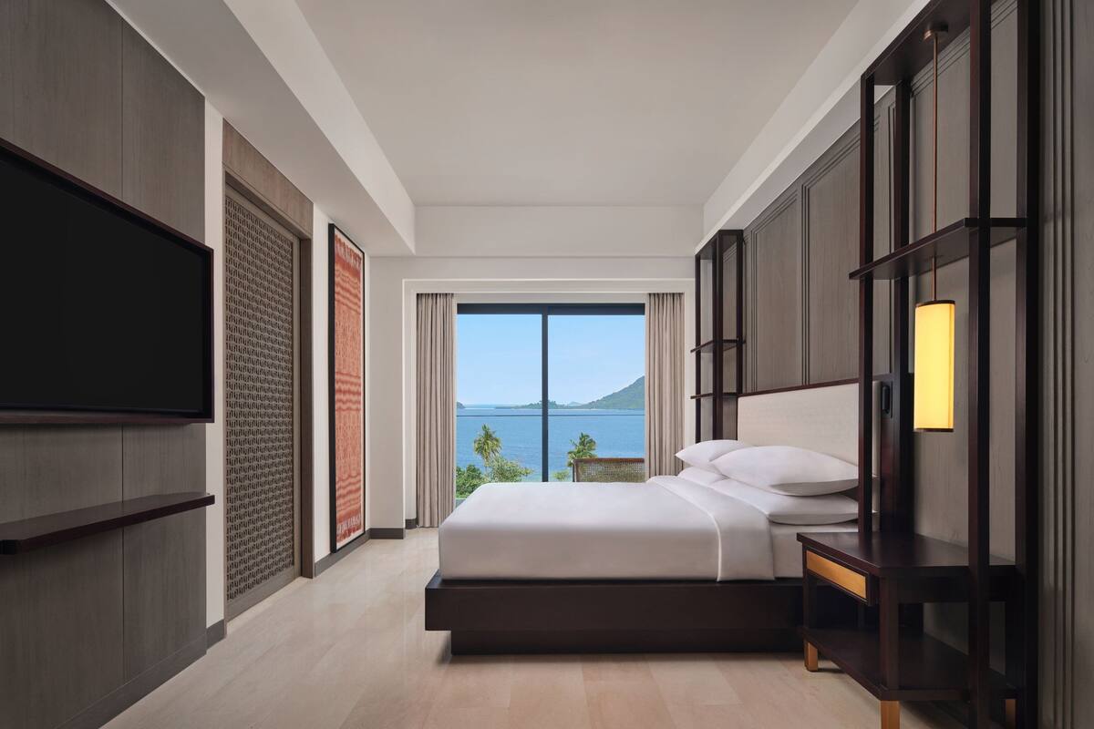 A modern hotel room with a large bed, a TV, and a view of the sea.