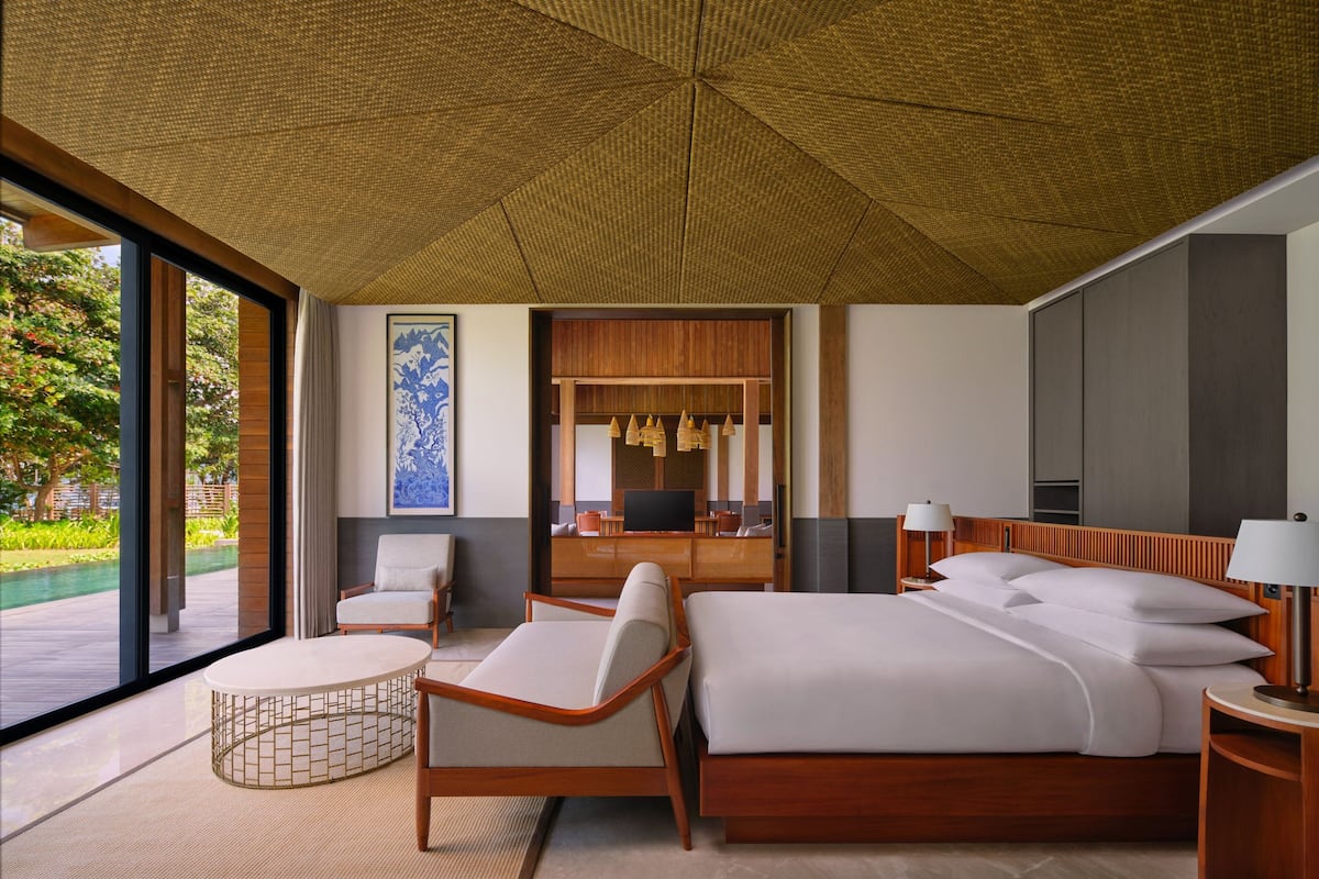 A modern hotel room with a large bed, two armchairs, a small round table, a TV, and a large window offering a view of a garden.