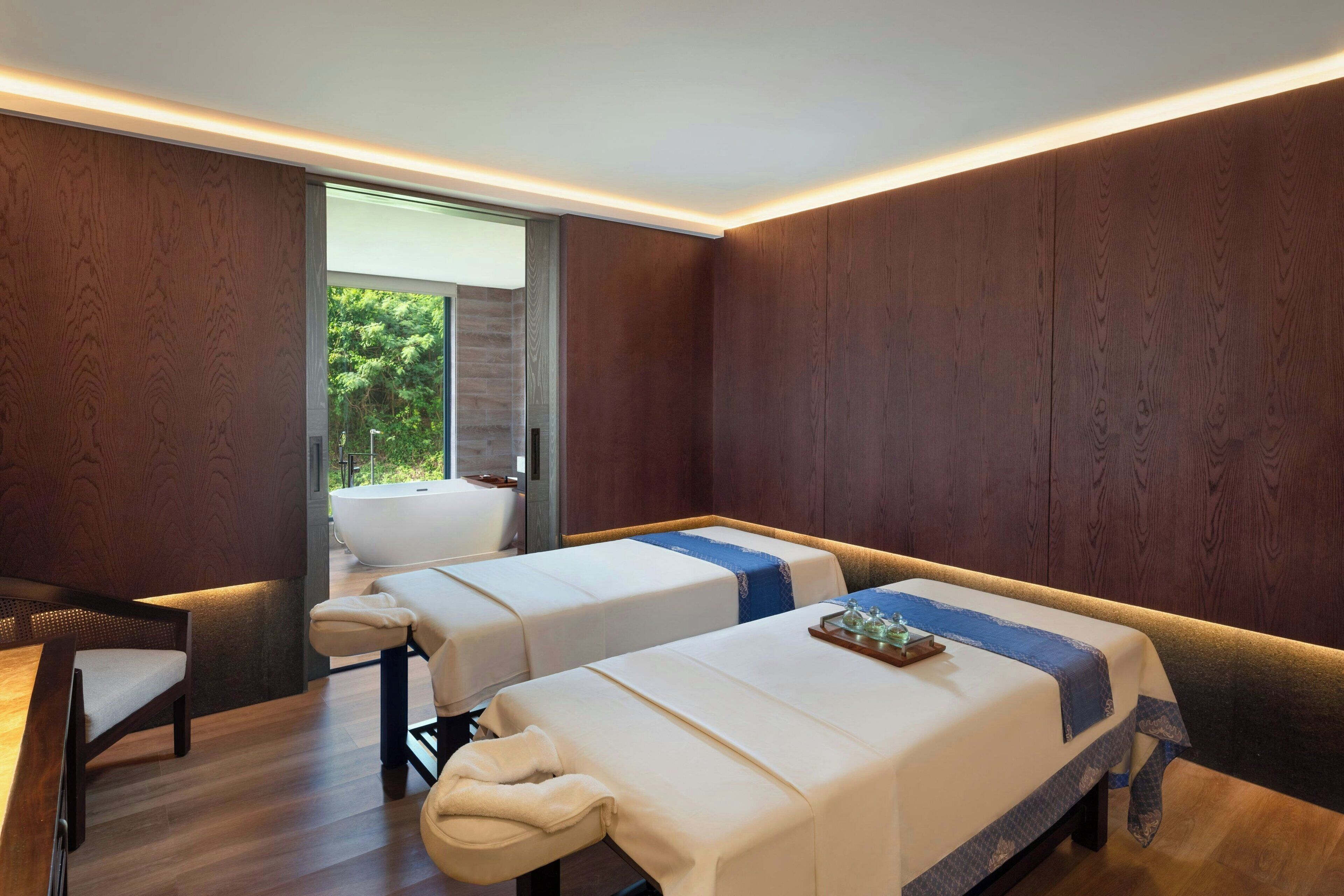 A spa room with two massage tables, a wooden bench, and a view of the outdoors.