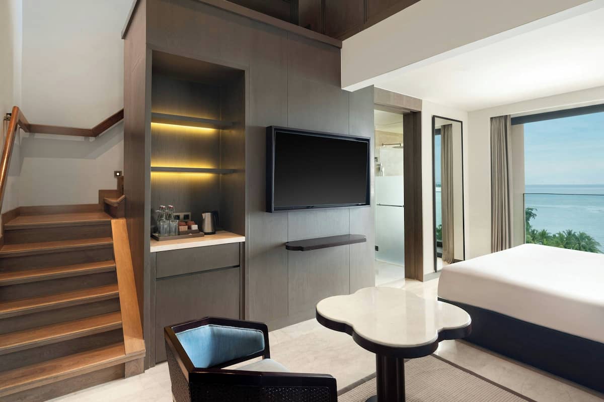 A modern hotel room with a large bed, a TV mounted on the wall, a small table, and a staircase leading to a balcony with a view of the sea.