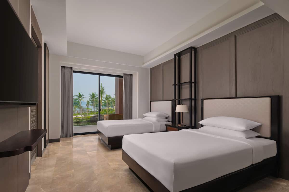 A hotel room with two beds, a TV, a desk, and a balcony with a view of palm trees.