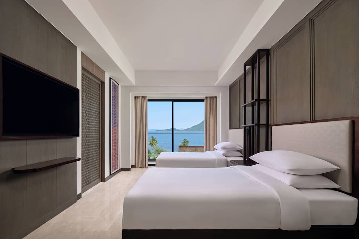 A hotel room with two beds, a TV, and a large window offering a view of the sea.
