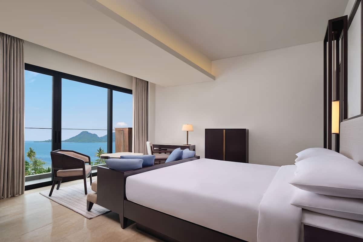 A modern hotel room with a large bed, a desk, a chair, and a view of the ocean.