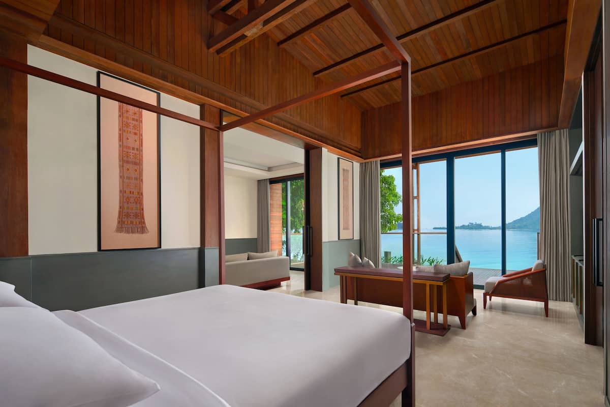 A modern hotel room with a large bed, a sitting area with a table and chairs, and a view of the sea.