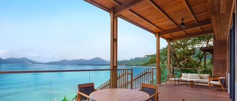 A wooden deck with a round table and chairs, overlooking a clear blue sea and distant mountains.