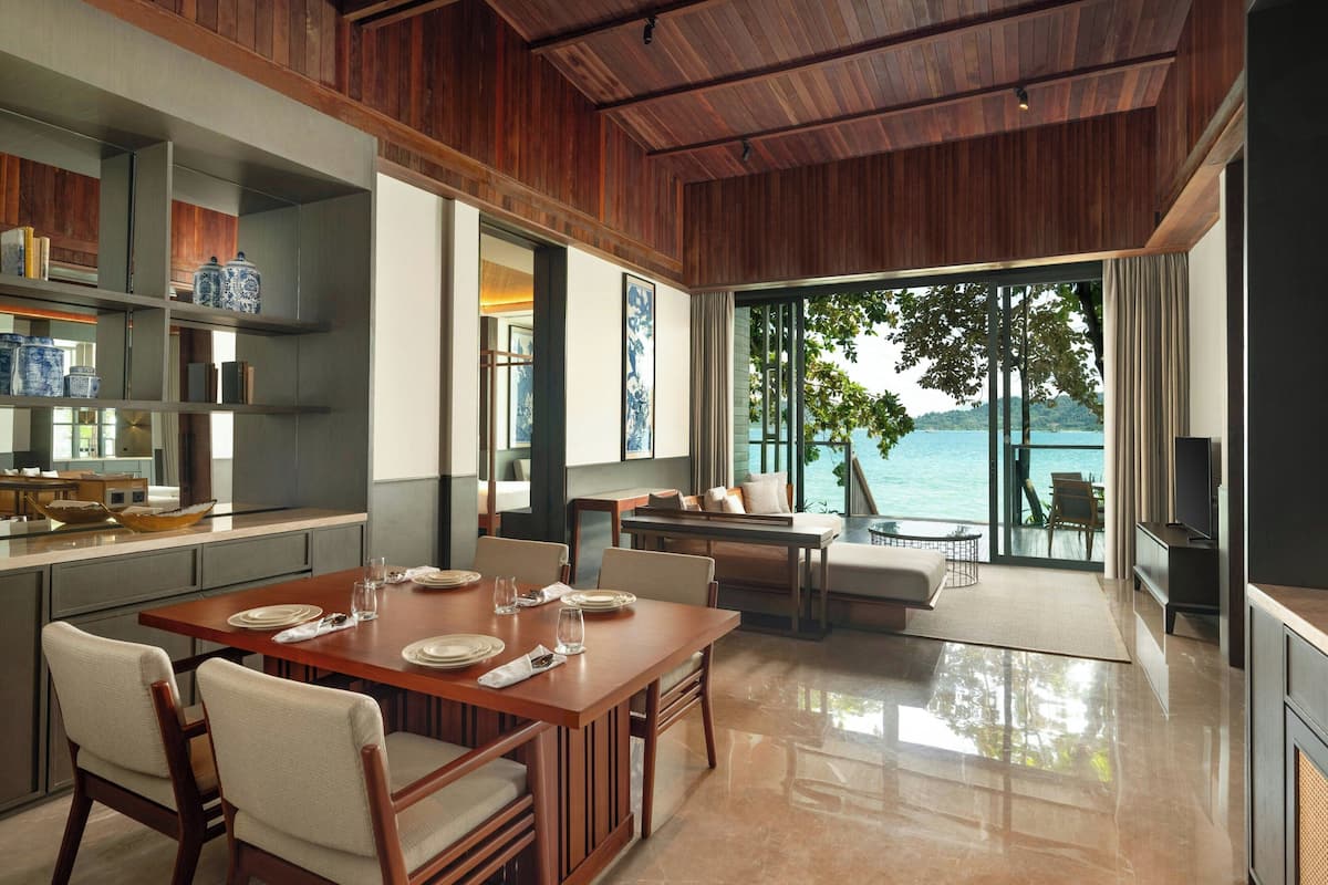 A modern dining area with a wooden table set for four, a kitchen with open shelving, and a view of the sea through large windows.