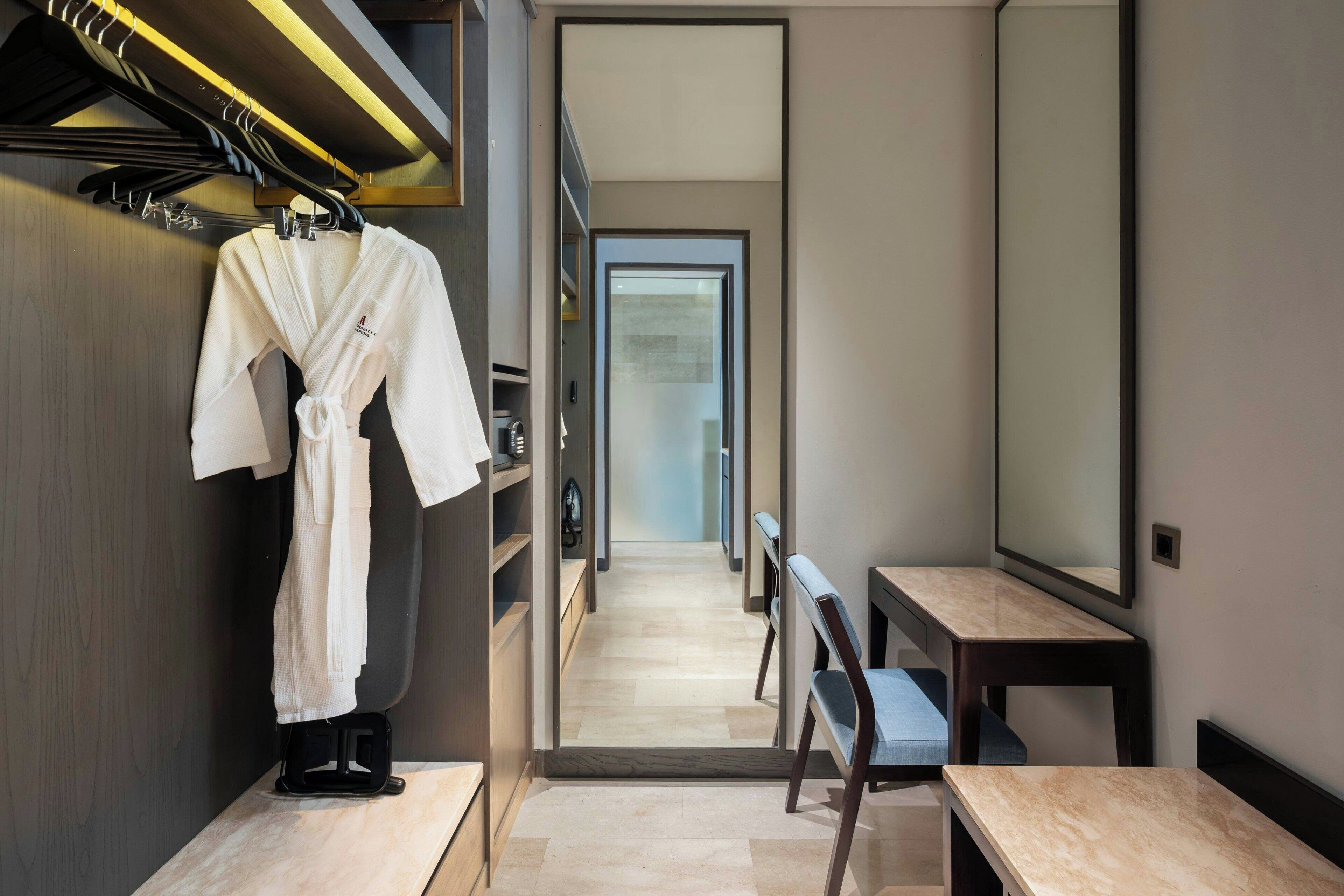A modern hotel room with a robe hanging on a rack, a mirror, and a wooden bench.