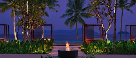 A fire pit surrounded by lush greenery and palm trees, with a view of the ocean at dusk.