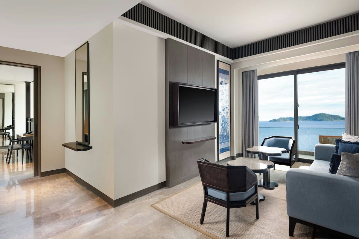 A modern living room with a flat-screen TV, a sofa, a dining area, and a view of the water.