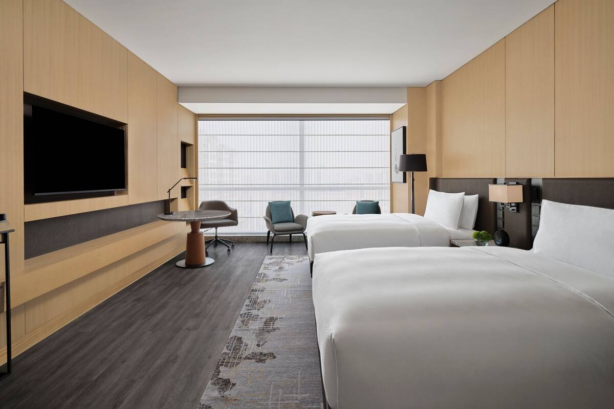 Een hotelkamer met twee bedden, een bureau, een stoel, een televisie en een groot raam met jaloezieën.