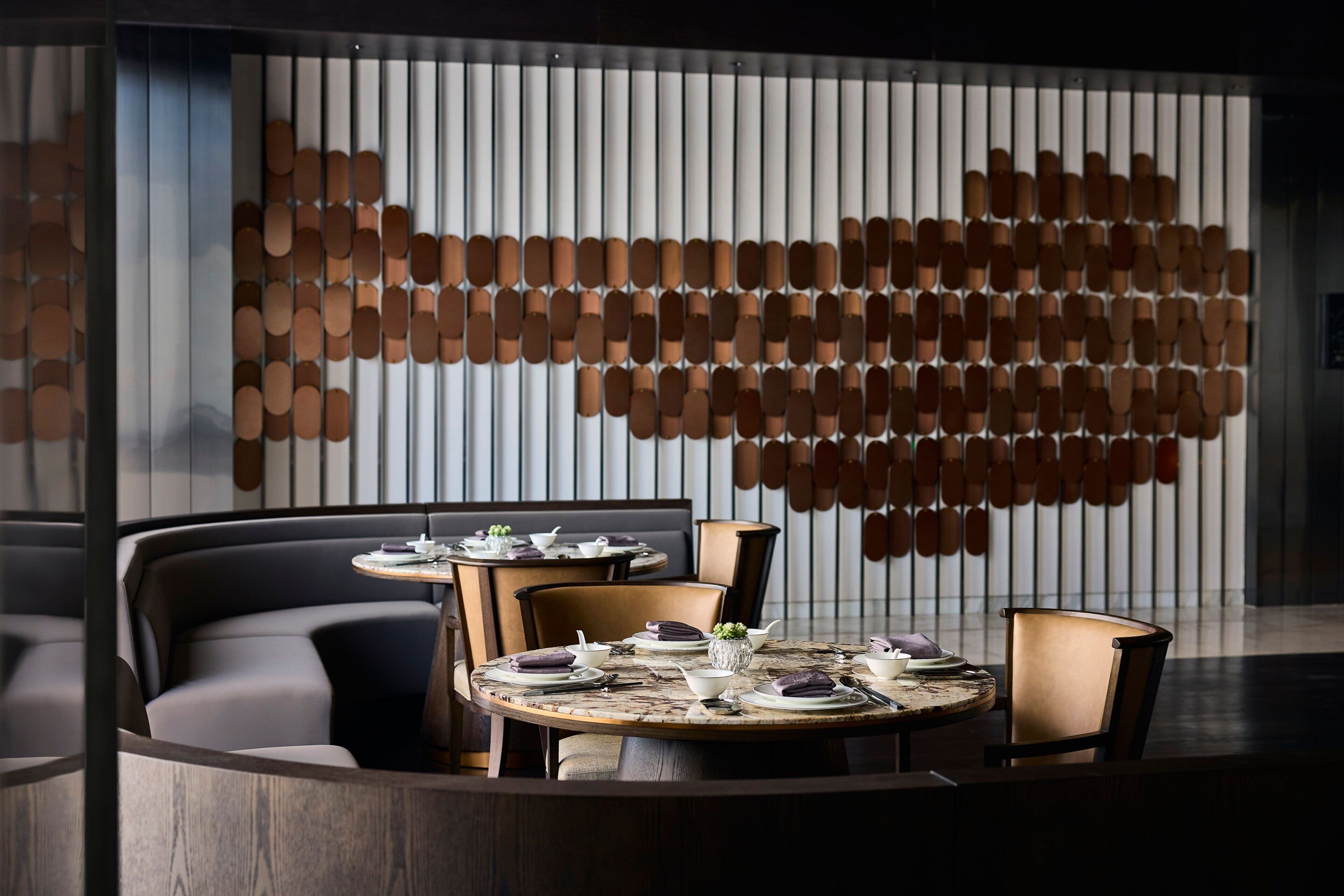 Een moderne eetruimte met ronde tafels en zachte zitplaatsen, een decoratieve wand met een patroon van cirkelvormen en een neutraal kleurenpalet.