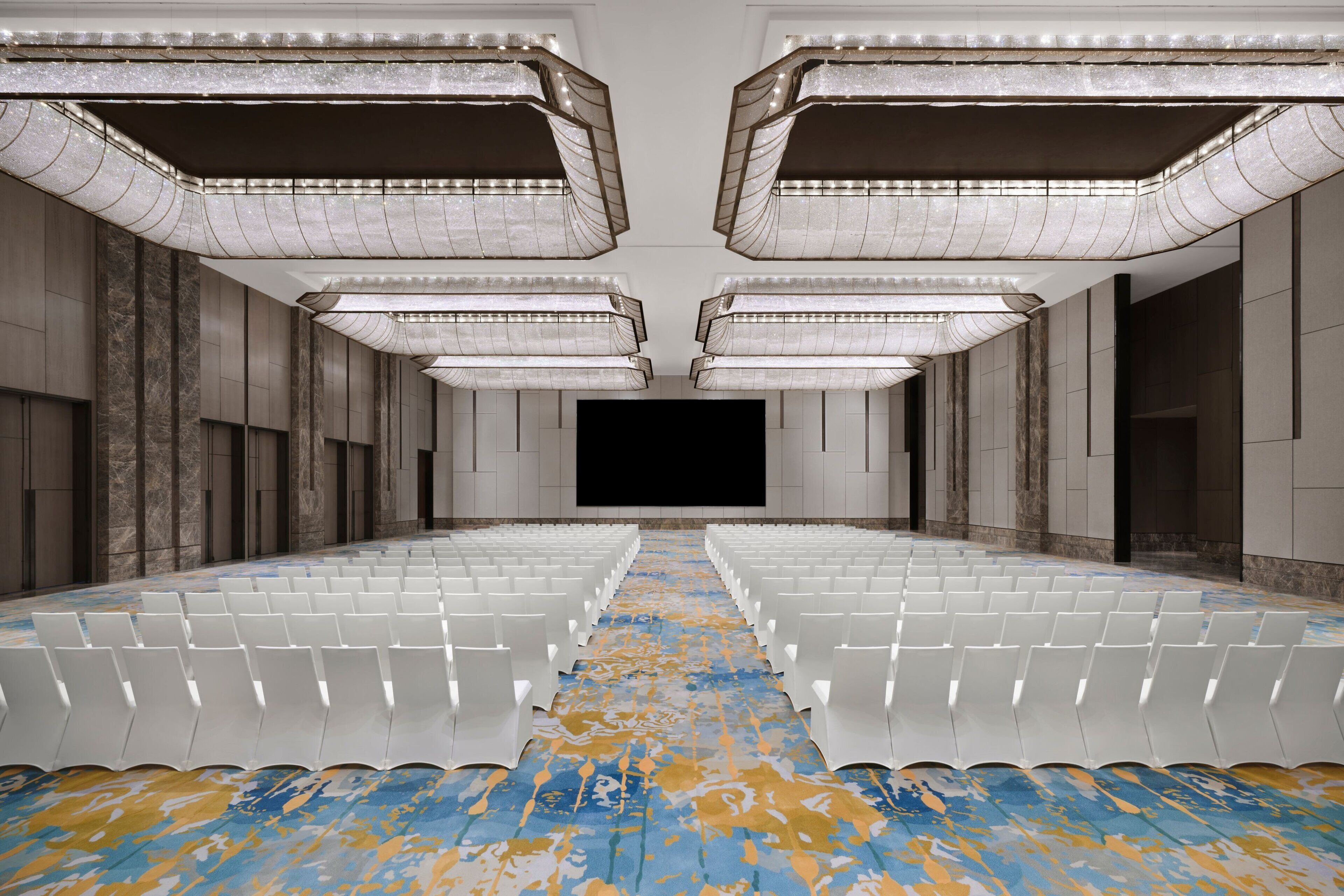 Un grand hall vide, avec des rangées de chaises blanches, un sol à motifs et un écran noir au centre.