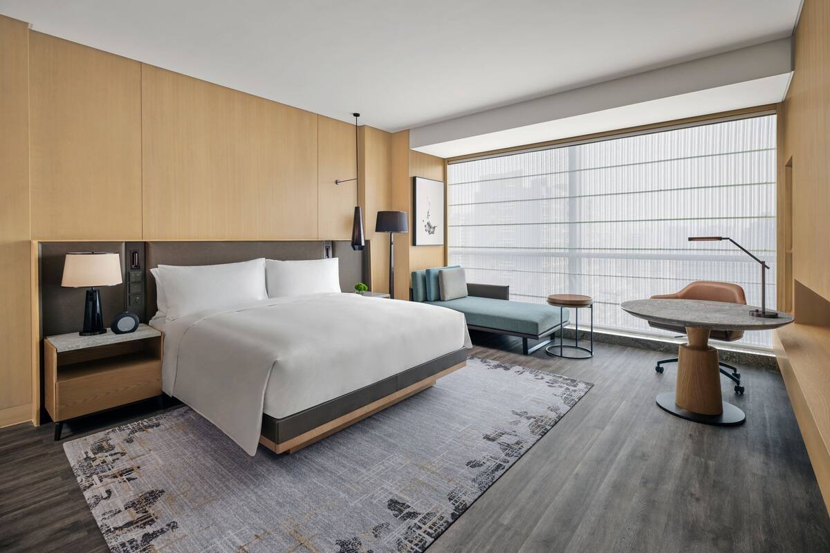 Een moderne hotelkamer met een groot bed, een bureau, een stoel en uitzicht op de stad door grote ramen.