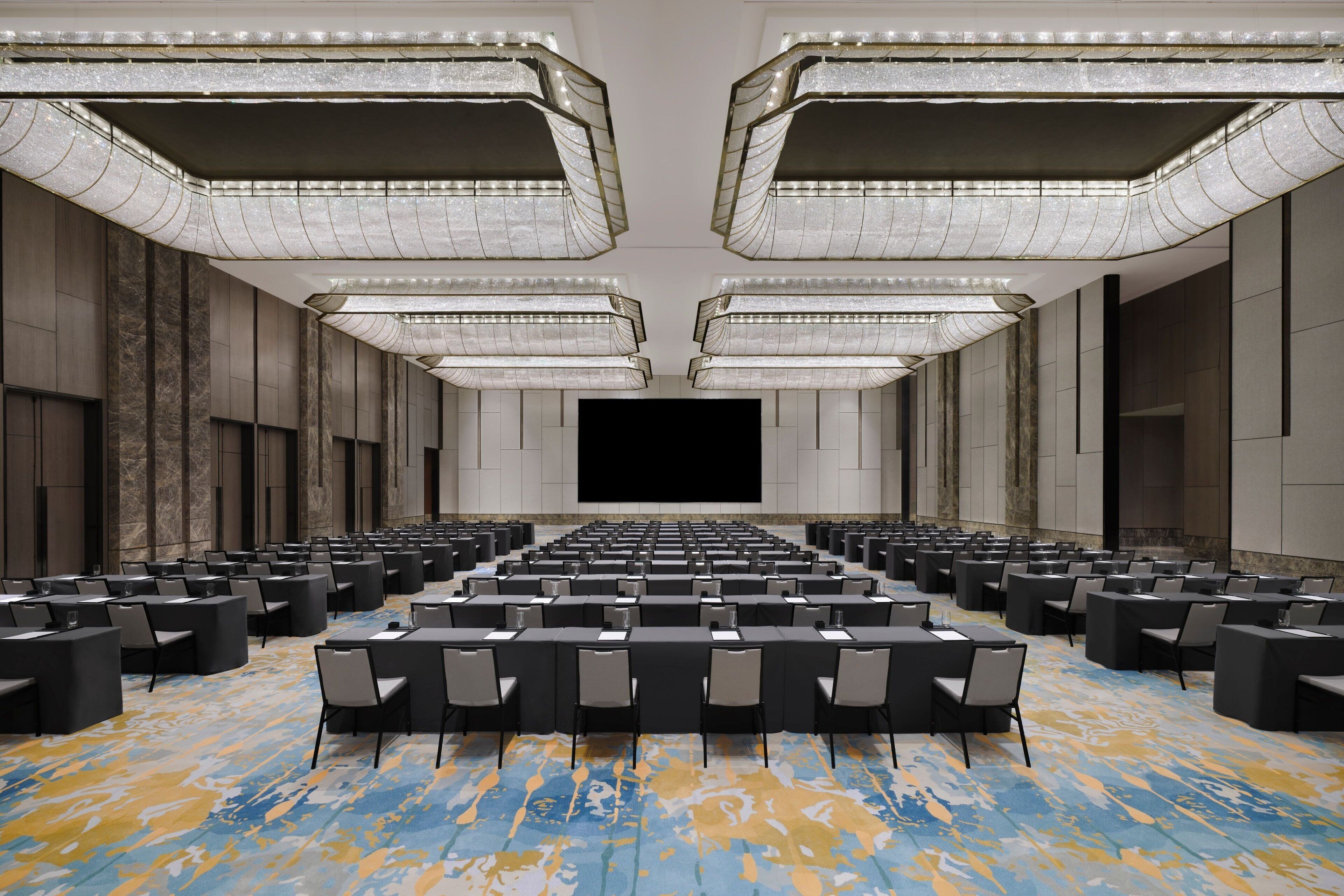 A conference room with rows of tables and chairs, a large screen, and a patterned carpet.
