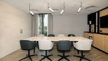 A conference room with a long table, four chairs, a white brick wall, a large window with curtains, and a kitchenette area.