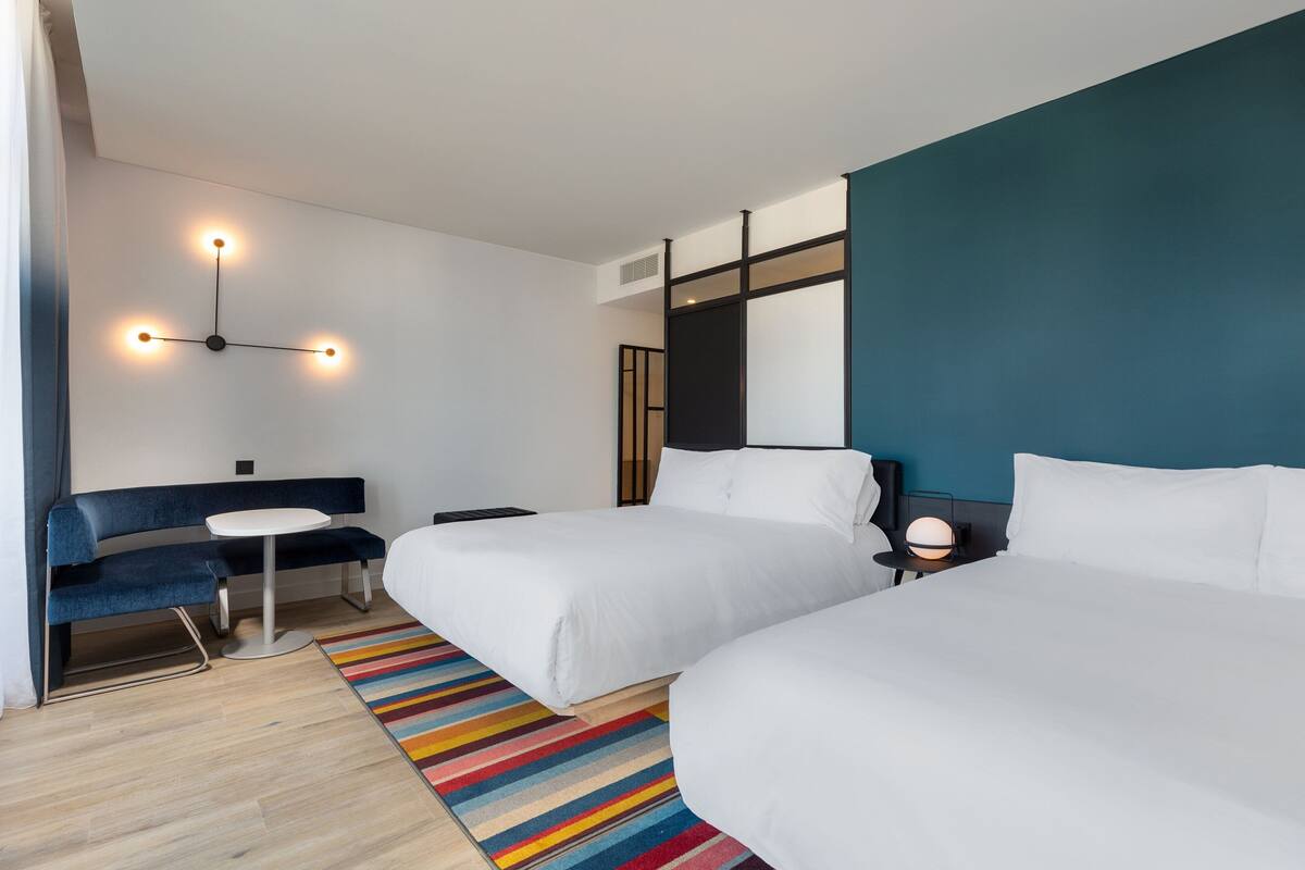 Ein Hotelzimmer mit zwei Betten, einem kleinen runden Tisch, einer Couch, einem bunten Streifen Teppich und einer modernen Lampe.
