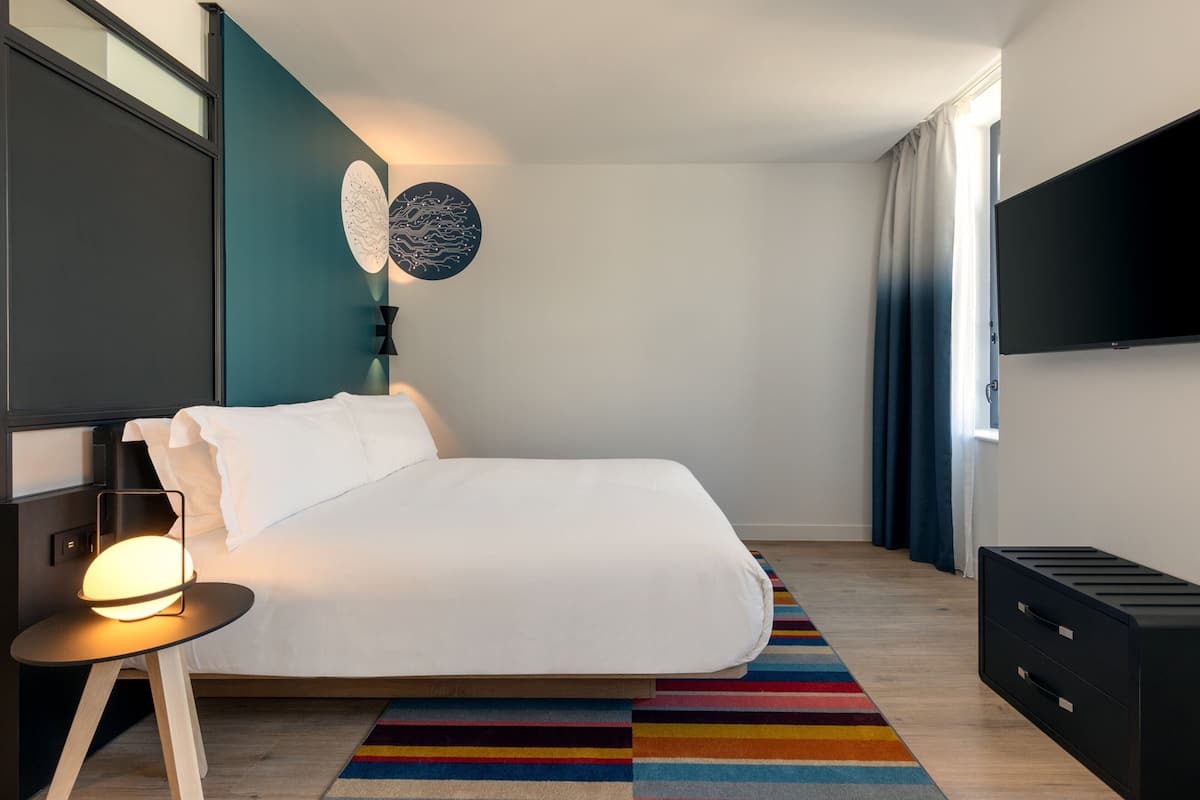 Ein modernes Hotelzimmer mit einem großen Bett, einem Nachttisch mit Lampe, einem an der Wand montierten Flachbildfernseher und einem farbenfrohen, gestreiften Teppich.