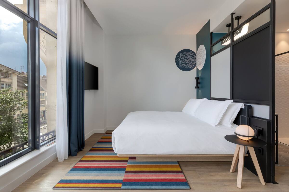 Un dormitorio moderno con un ventanal grande, una cama, un tapete a rayas de colores y un televisor montado en la pared.