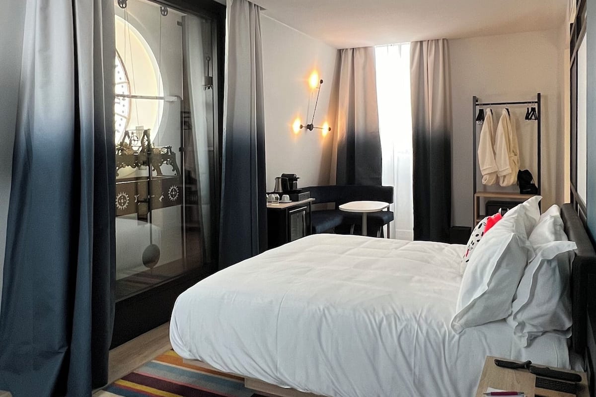 Una habitación de hotel moderna con una cama grande, un escritorio y una silla. Hay una ventana con cortinas, una lámpara de pared y un perchero con ropa.