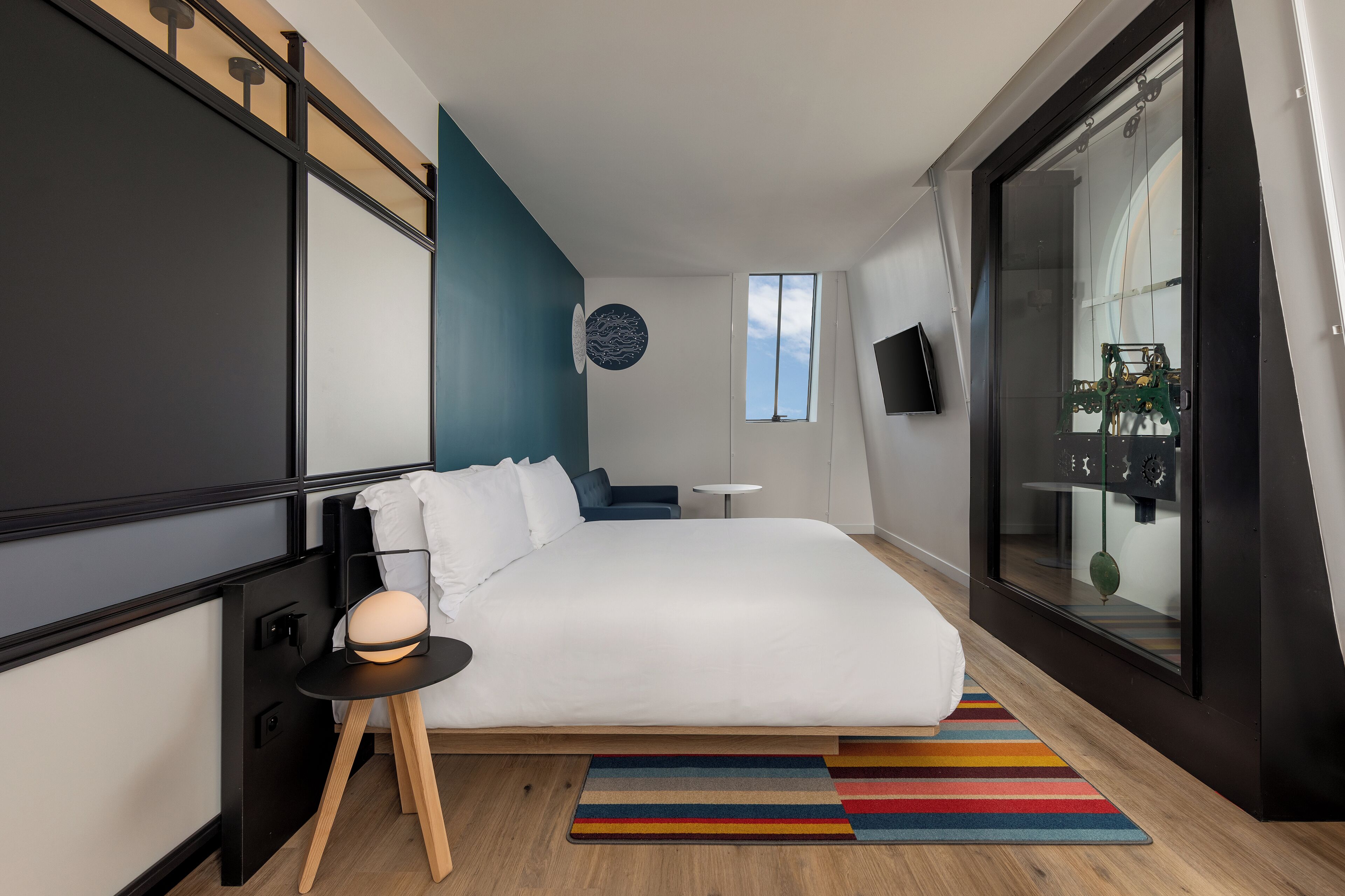 Una habitación de hotel moderna con una cama grande, una mesita auxiliar pequeña, una alfombra colorida a rayas, un televisor montado en la pared y un ventanal con vistas al cielo.