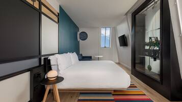 Una habitación de hotel moderna con una cama grande, una mesita auxiliar pequeña, una alfombra colorida a rayas, un televisor montado en la pared y un ventanal con vistas al cielo.