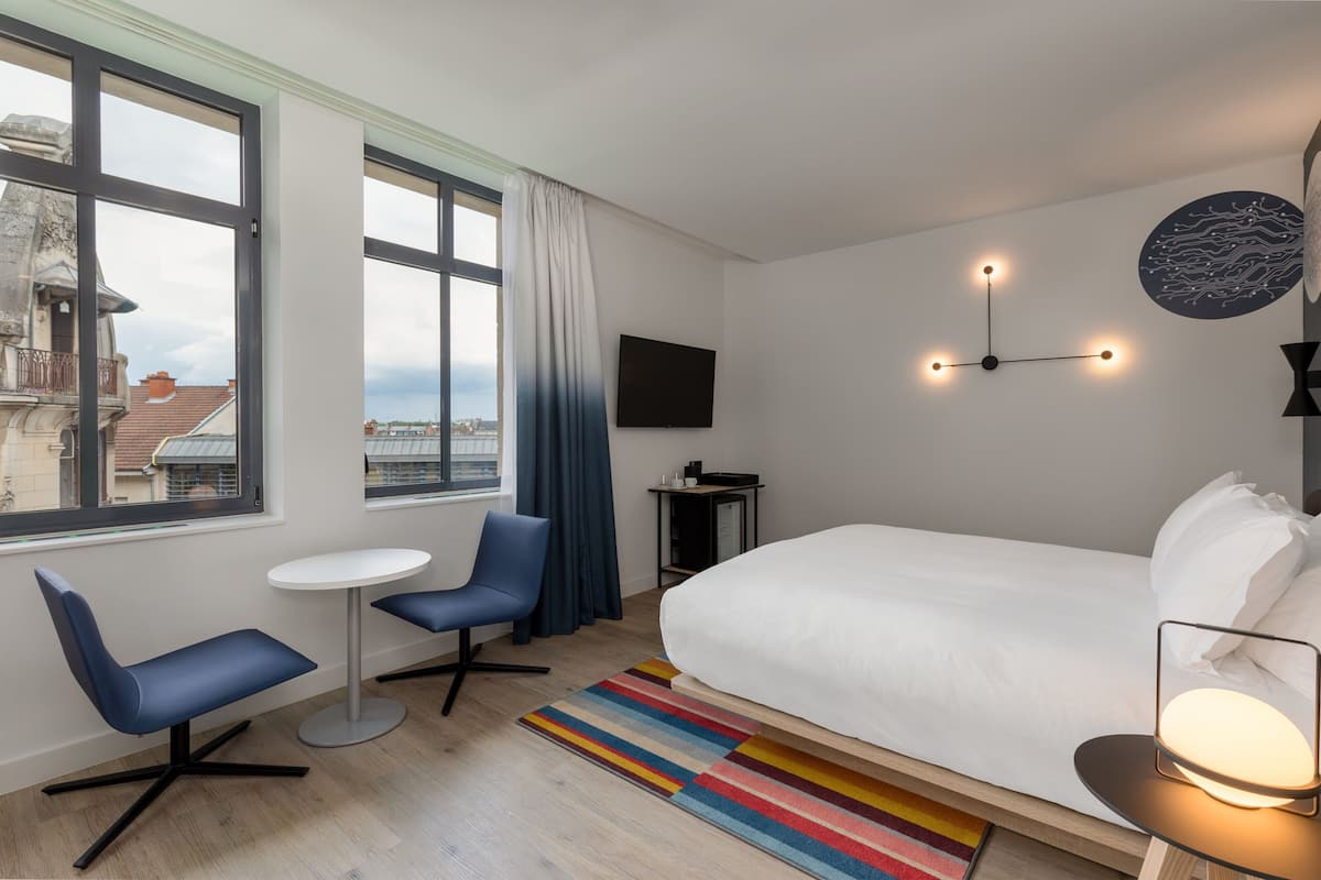 Un hotel moderno con una cama grande, una mesita redonda pequeña, una silla azul, un televisor montado en la pared y vistas de la ciudad a través de amplios ventanales.