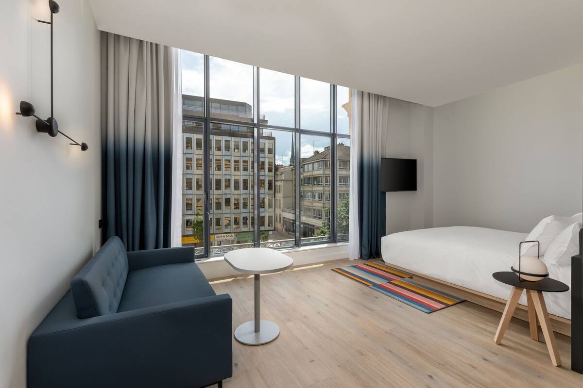 Ein modernes Hotelzimmer mit großem Fenster, einer Couch, einem Bett, einem kleinen Tisch und einem Fernseher.
