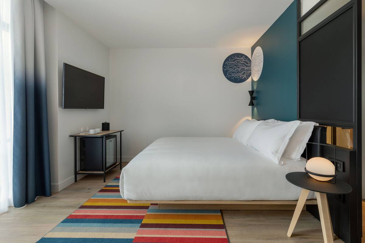 Ein modernes Schlafzimmer mit einem großen Bett, einem Nachttisch, einem an der Wand montierten Fernseher und einem farbenfrohen Streifen Teppich.