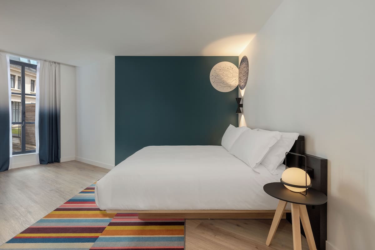 Un dormitorio moderno con una cama grande, un tapete a rayas de colores, una lámpara de pared y vistas al exterior.