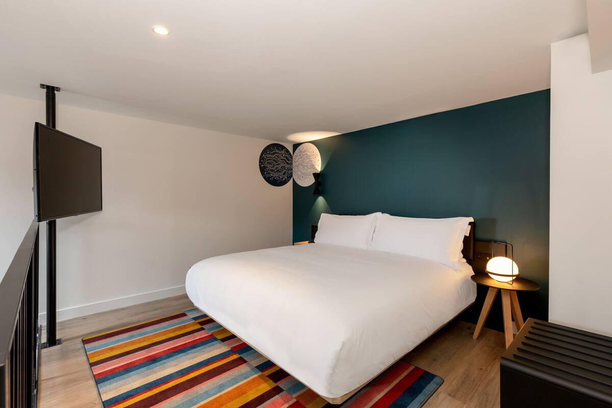 Ein modernes Schlafzimmer mit einem großen Bett, einem farbenfrohen Streifen Teppich, einem an der Wand montierten Fernseher und einem Nachttisch mit einer Lampe.