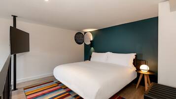 Une chambre moderne avec un grand lit, un tapis rayé aux couleurs vives, un téléviseur fixé au mur et une table de chevet avec une lampe.