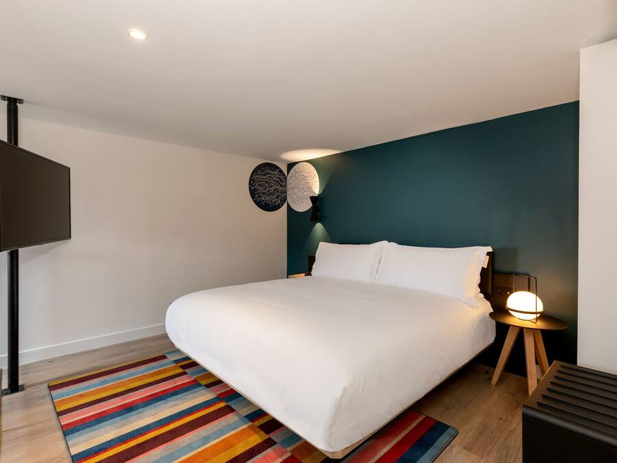 Une chambre moderne avec un grand lit, un tapis rayé aux couleurs vives, un téléviseur fixé au mur et une table de chevet avec une lampe.