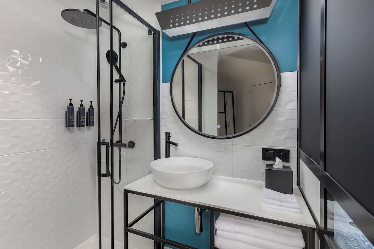 Un baño moderno con un lavamanos blanco, un espejo redondo y una ducha con cerramiento de vidrio.