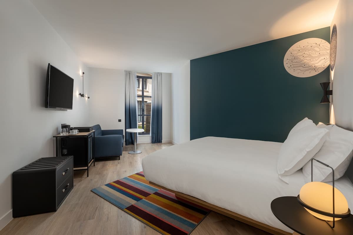 Ein modernes Hotelzimmer mit einem großen Bett, einem Schreibtisch mit Stuhl, einem Flachbildfernseher und einer kleinen Küchenzeile.