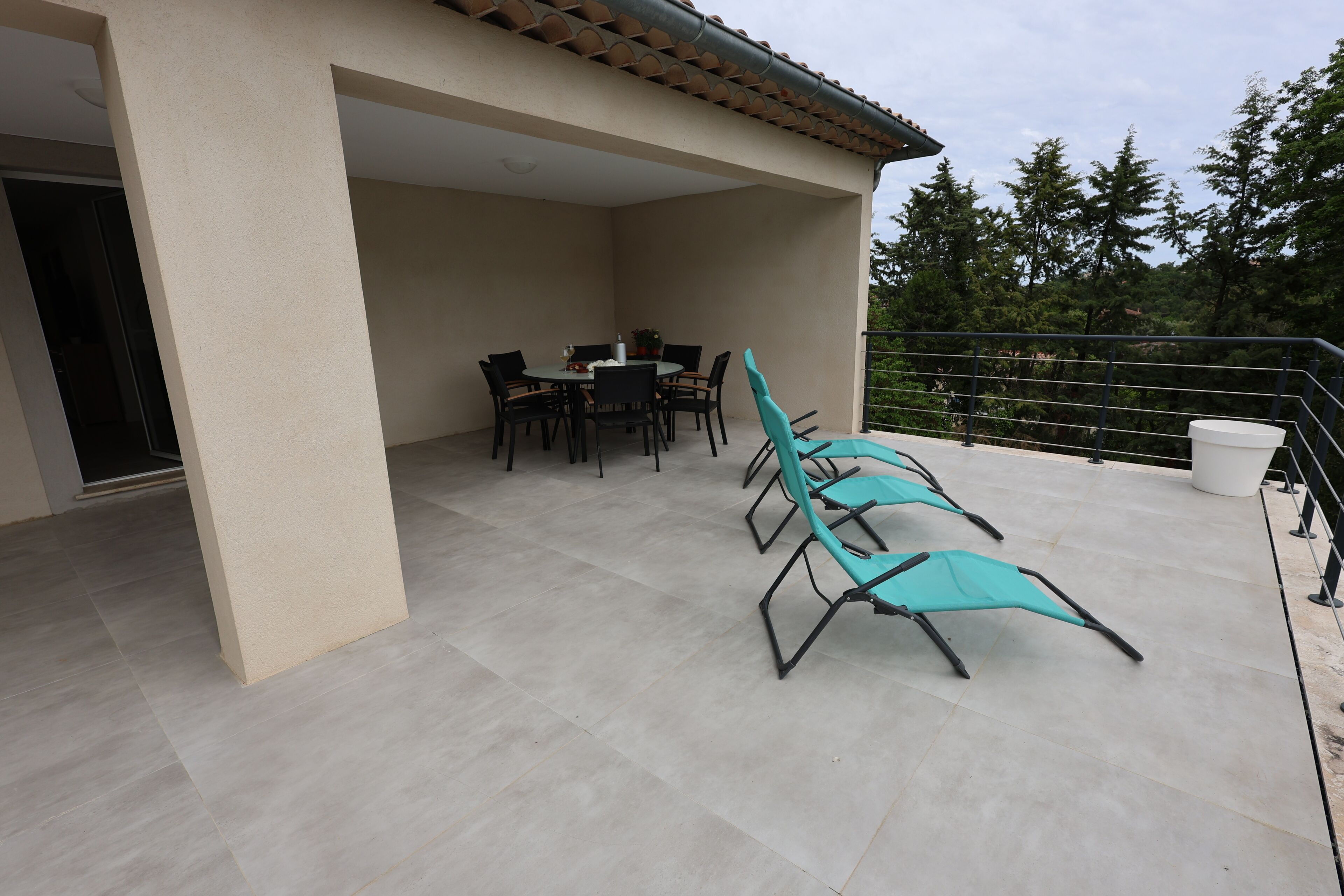 An outdoor patio area with a table and chairs, a covered seating area with lounge chairs, and a view of trees and a building in the background.