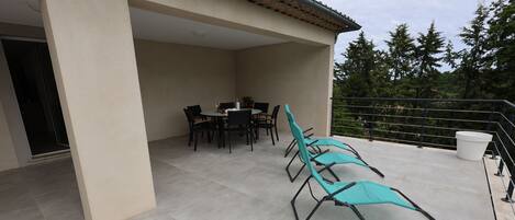 Un patio al aire libre con una mesa y sillas, un espacio de estar cubierto con sillones de descanso, y vista a árboles y un edificio al fondo.