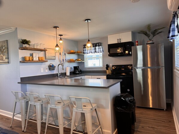 A modern kitchen with a central island, bar stools, stainless steel appliances, and decorative lighting. - Comfy Coastal Cottage One (Surfside Beach)