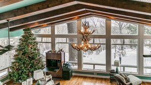 A spacious room with a large wooden ceiling, a fireplace, a Christmas tree, and a view of a snowy landscape through large windows. - Huge Retreat with Heated Pool And Hot Tub by Briggs Woods Walking Trail and Golf (Webster City)