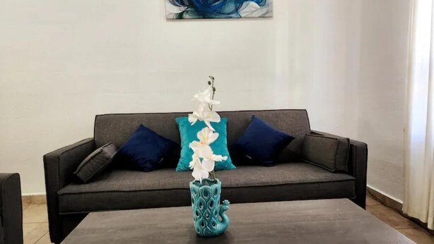 Un salon avec un canapé gris foncé, des coussins turquoise et bleus, un vase turquoise orné de fleurs blanches et un tableau abstrait bleu accroché au mur.