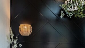Ein Schlafzimmer mit schwarzer geometrischer Wand, einer gewebten Leuchte, einer hängenden Pflanze, einem Nachttisch mit Büchern und einem Bett mit weißer Bettwäsche.