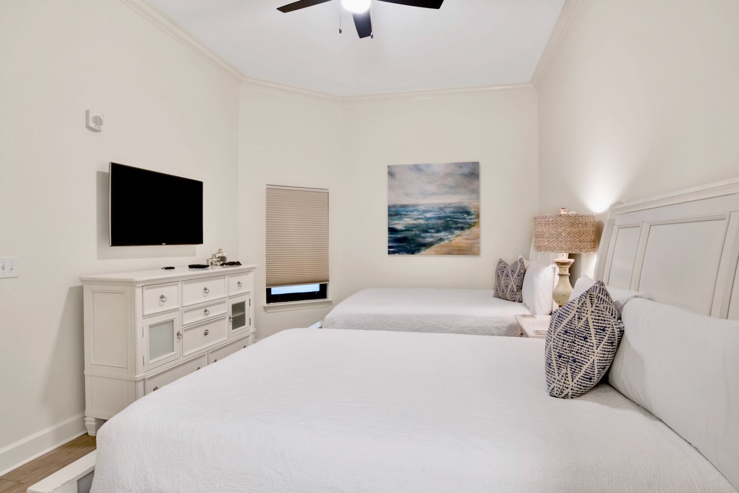 A bedroom with two beds, a TV, a dresser, a painting, and a ceiling fan.
